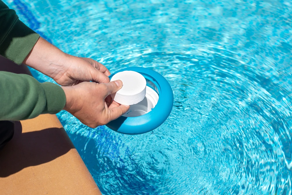 Gros plan sur une main tenant un kit d'analyse de l'eau sur une piscine claire et bleue, avec un équipement de nettoyage de piscine visible en arrière-plan