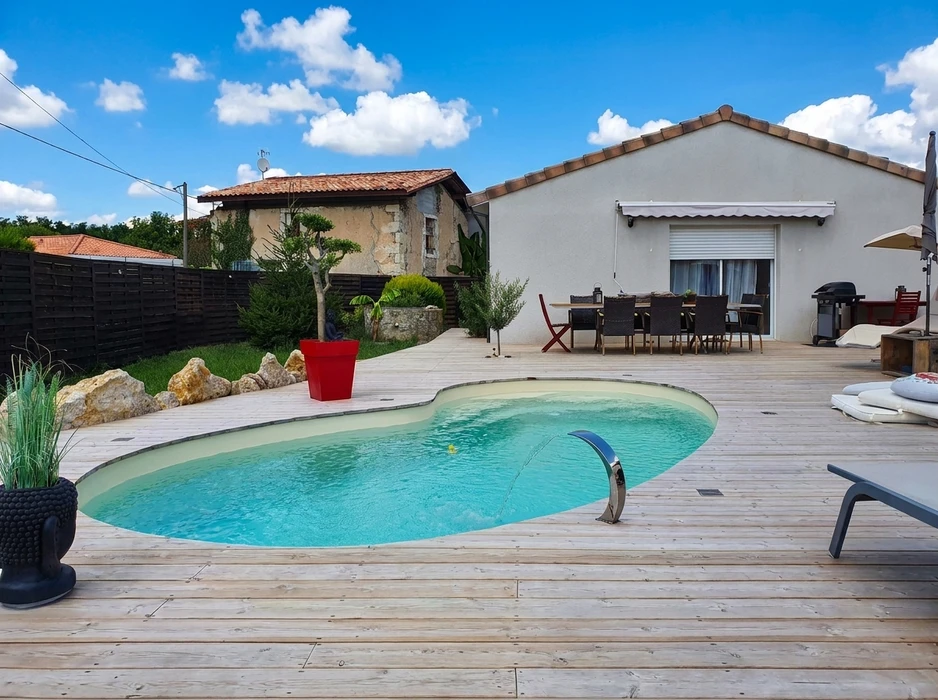 Photographie professionnelle d'une piscine résidentielle haut de gamme en béton armé, photographiée en fin d'après-midi avec un éclairage doré. La piscine présente des lignes épurées et modernes, avec une eau cristalline d'un bleu profond et lumineux. Elle est équipée d'un système de traitement visible (débordement intégré ou cascade d'eau). Une large plage en dalle de pierre naturelle l'entoure, avec quelques transats de luxe et un parasol. Un aménagement paysager sophistiqué (arbustes taillés, massifs de fleurs, éclairage paysager subtil) encadre la scène. Un coin lounge avec mobilier haut de gamme est visible à proximité. La photographie, prise de jour en conditions optimales, doit mettre en évidence le caractère haut de gamme, la finition professionnelle et le prestige de cette installation complète clé en main, distinct des trois images existantes par son focus sur le luxe et la sophistication du projet terminé.
