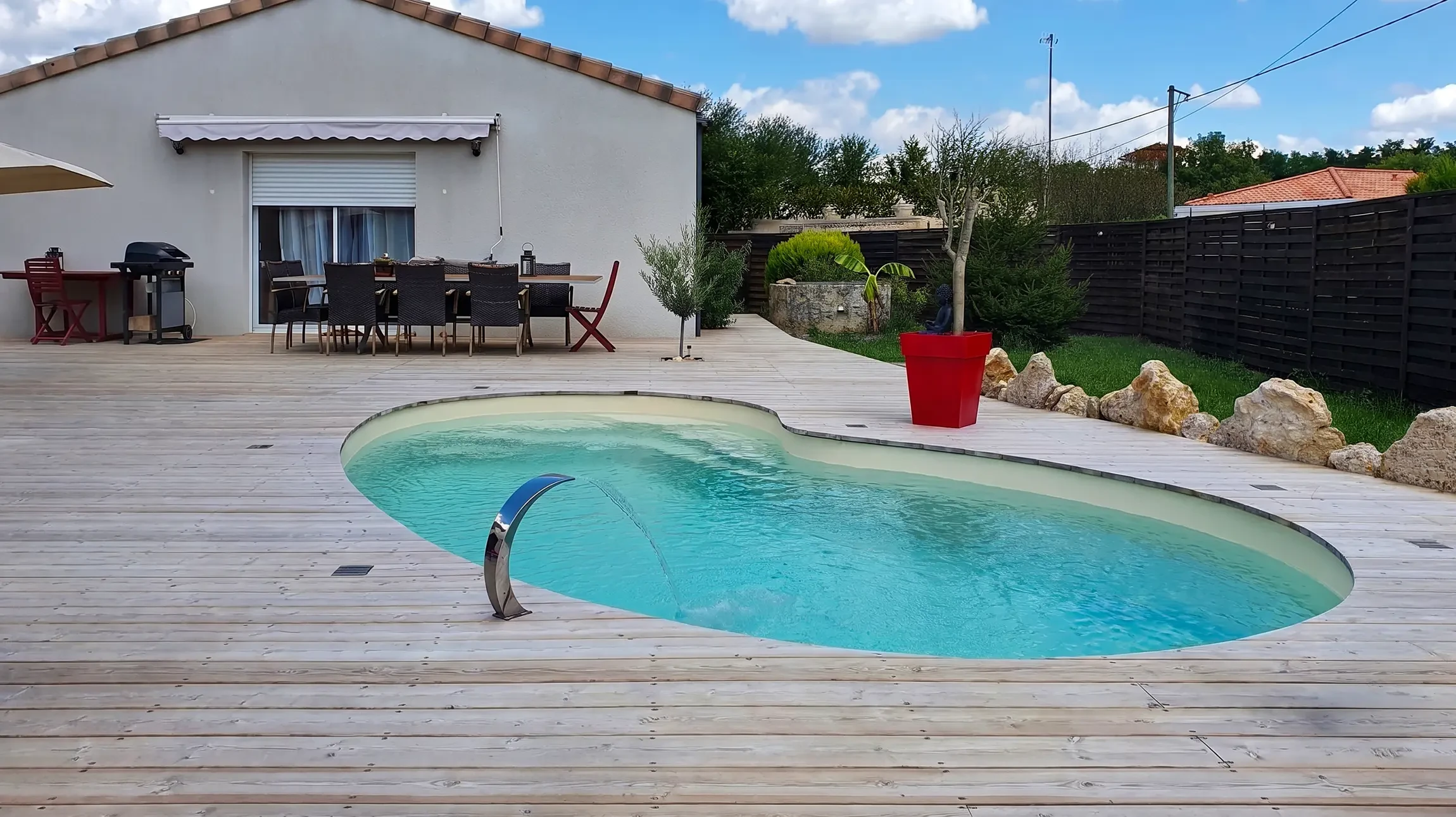 Photographie réelle d'une piscine coque polyester de forme moderne installée dans un jardin aménagé, vue d'angle montrant la piscine remplie d'eau claire bleue turquoise, entourée d'une terrasse en bois ou dallage, avec pelouse verte et arbustes en arrière-plan, sous un ciel dégagé. Pas de personnage visible. La piscine doit être clairement reconnaissable comme une coque polyester lisse de qualité, illustrant un aménagement complet et professionnel dans un cadre résidentiel.